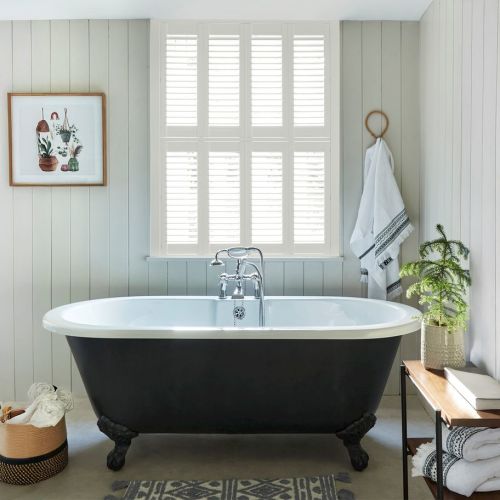 White plantation shutters (six-panel, horizontal slats) cover the central window, filtering bright daylight into a pale paneled bathroom with a black clawfoot tub, towel, plant, and bench.