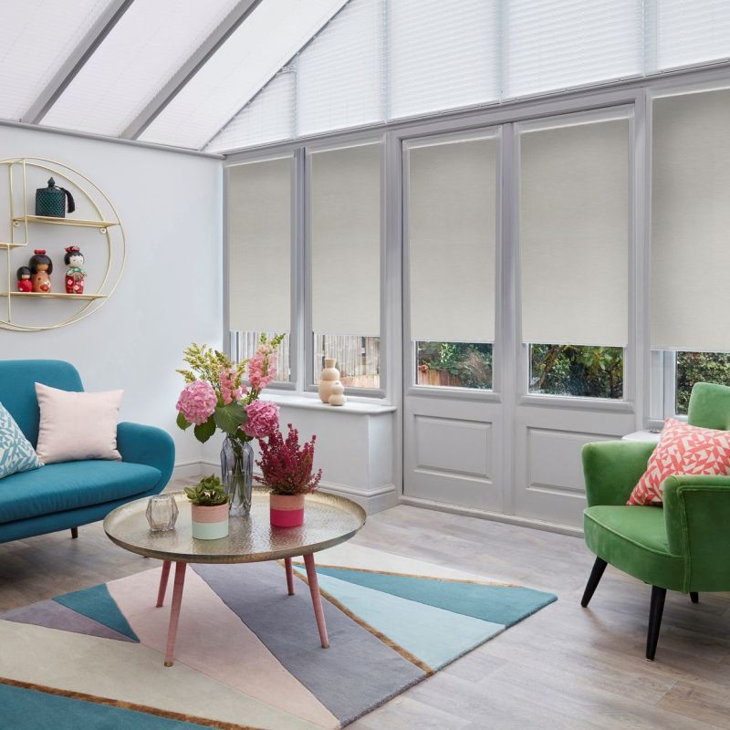 Beige roller blinds, half-lowered over tall glazed doors and windows, filtering daylight; conservatory-style lounge with teal sofa, green armchair, round coffee table and flowers on a pastel geometric rug.