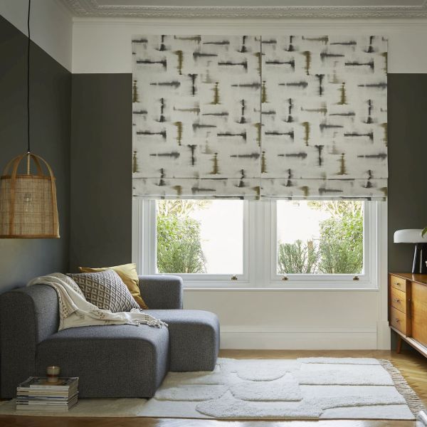 Patterned fabric Roman blinds—paired over twin windows with abstract gray‑olive brushstroke print—are partially lowered, softly diffusing daylight; cozy living room with gray chaise, woven pendant lamp, wooden sideboard, garden view.