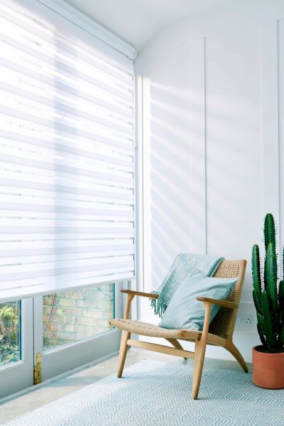 White zebra roller blind over glass door, alternating sheer and opaque stripes, lowered and filtering bright daylight; rattan chair with mint cushion and throw, potted cactus in sunlit room.