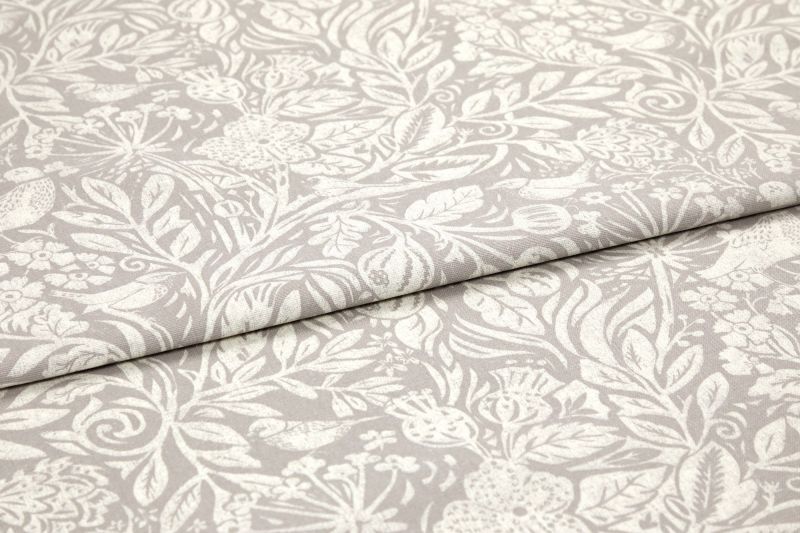 Folded beige cotton-linen fabric with white botanical print, overlapping fold across center; pattern of leaves, flowers and birds; close-up on a flat surface with soft, even lighting.