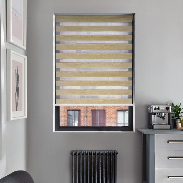 Zebra-style roller blind with alternating beige wood‑grain and sheer horizontal stripes, partially lowered, filtering daylight; set in a grey-walled kitchen nook with a coffee machine, radiator below, and brick building view.