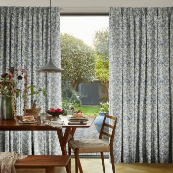 Full-length pleated curtains with blue floral print, pulled aside to frame an open gap; filtering daylight into a dining area with wooden table set for breakfast and a backyard.