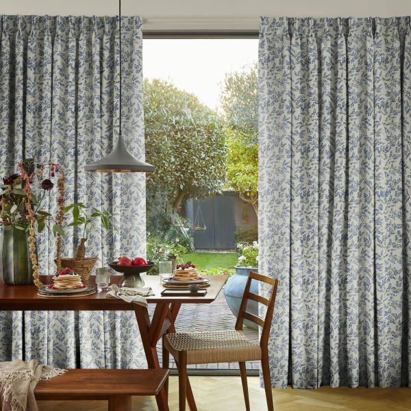 Full-length pleated curtains with blue floral print, pulled aside to frame an open gap; filtering daylight into a dining area with wooden table set for breakfast and a backyard.