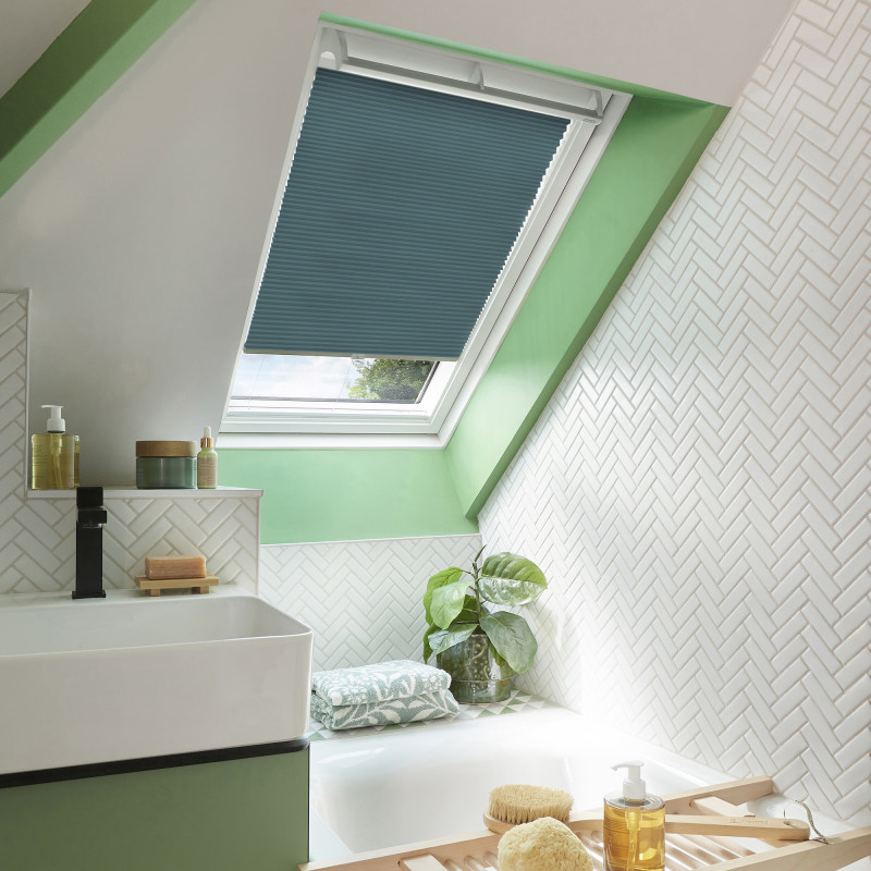 Teal pleated skylight blind, almost fully lowered over a roof window, filtering daylight; attic bathroom setting with green-painted window recess, white herringbone tiles, sink and bathtub with plant and toiletries.