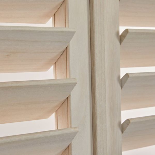 Light unfinished wood plantation shutters with angled horizontal louvers and a central stile — louvers tilted open, filtering soft daylight — close-up interior view of adjacent panels.