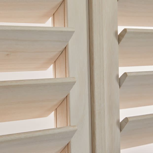 Light unfinished wood plantation shutters with angled horizontal louvers and a central stile â€” louvers tilted open, filtering soft daylight â€” close-up interior view of adjacent panels.
