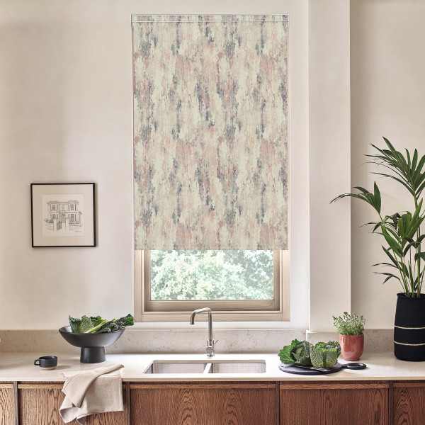 Patterned roller blind hangs down, partially covering a window above a kitchen sink; stainless faucet, wooden cabinets, potted plants and a framed sketch sit on the light countertop.