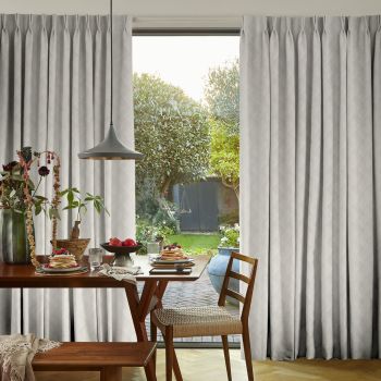 Floor-to-ceiling pleated light-gray curtains, parted centrally to reveal sliding glass door; curtains filter soft daylight. Interior: wooden dining table set for breakfast; view: sunlit garden with trimmed trees and lawn.