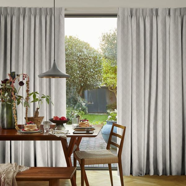 Floor-to-ceiling pleated light-gray curtains, parted centrally to reveal sliding glass door; curtains filter soft daylight. Interior: wooden dining table set for breakfast; view: sunlit garden with trimmed trees and lawn.