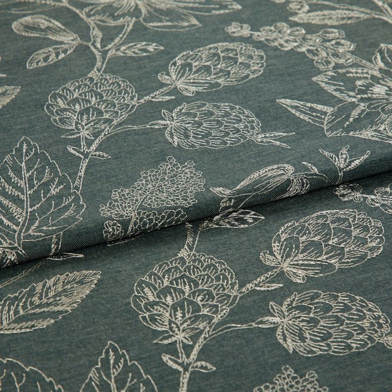 Dark teal fabric with white stitched botanical motifs (artichoke-like blooms and leaves), folded across the center, displaying layered, textured weave under soft, neutral studio lighting.