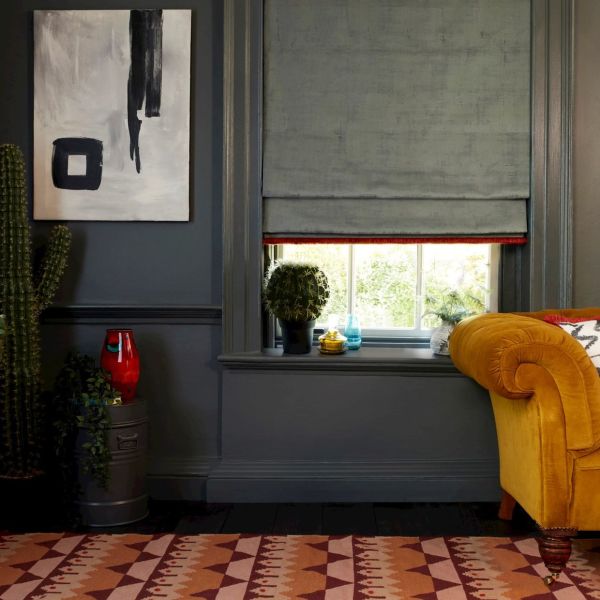 A gray Roman shade with red trim, partially lowered and filtering daylight through a sash window; in a dark-gray living room beside a mustard sofa, cactus and abstract painting.