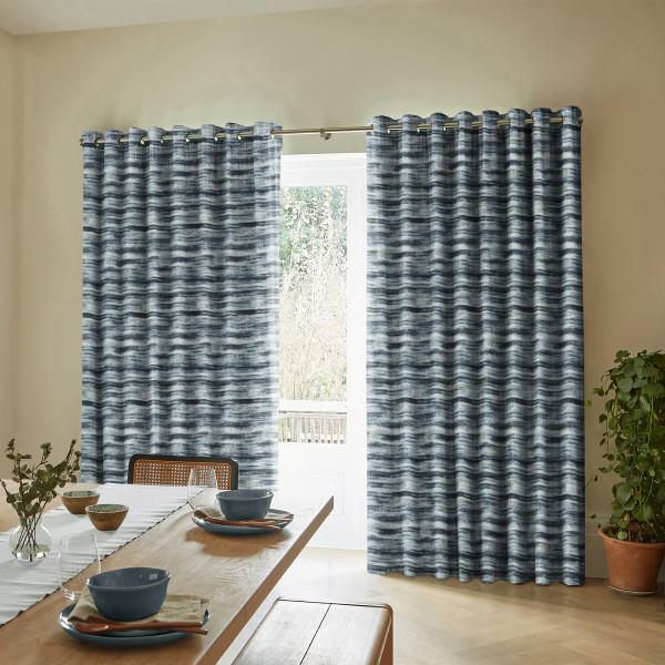 Blue-gray, horizontally striped grommet-top curtains hung on a metal rod, mostly closed with a central gap revealing a glass door; sunlit dining room with wooden table, bench, and potted plant.