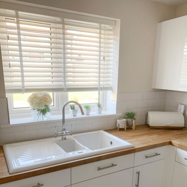 White horizontal blinds over the window, partially lowered with slats tilted to filter soft daylight, above a double white sink on wooden countertop in a bright kitchen with small plants.