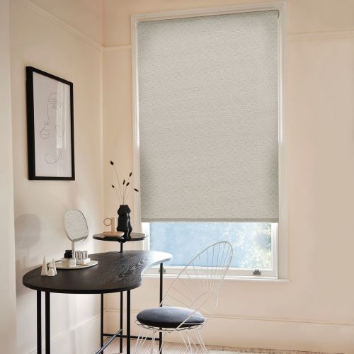 Light-gray textured roller blind, fully lowered with a narrow bottom gap, filtering soft daylight; set in a pale-walled minimalist home office with black curved desk, wire chair, and framed line-art.