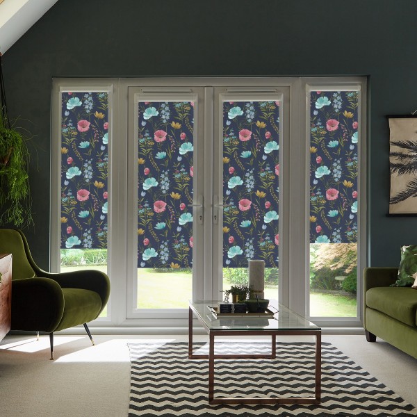 Floral-patterned roller blinds cover tall French doors and sidelights, blocking light in a modern living room with green armchairs, glass coffee table, chevron rug and glimpses of a garden.