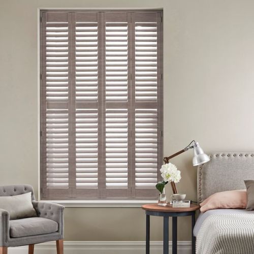 Grey wooden plantation shutters cover a tall window in four louvered panels; closed with slats angled to filter soft daylight into a neutral bedroom with armchair and bedside lamp.