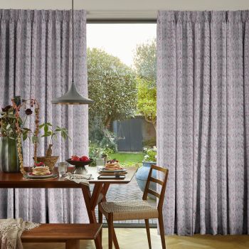 Floor-length pleated patterned curtains, pale grey with pink flecks, partially parted to frame a sliding glass door, filtering soft daylight over a breakfast table and lush garden view.