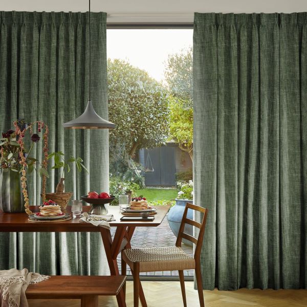 Floor-length, pleated green curtains hung on a ceiling track, partially drawn to frame a glass door; sunlit backyard visible beyond, dining table set with pancakes and wooden chairs inside.