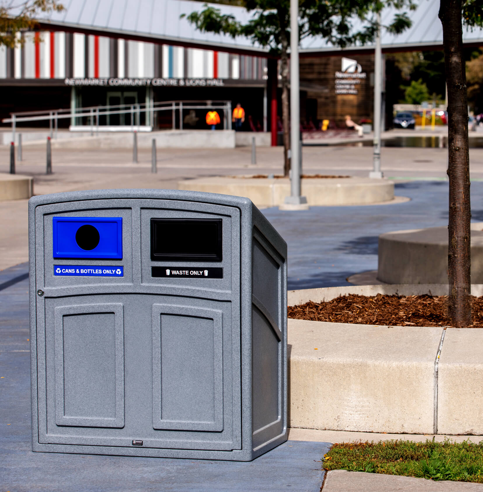 grey plastic recycling and waste bin with sloped top and front hinged door sitting outside at a municipal park