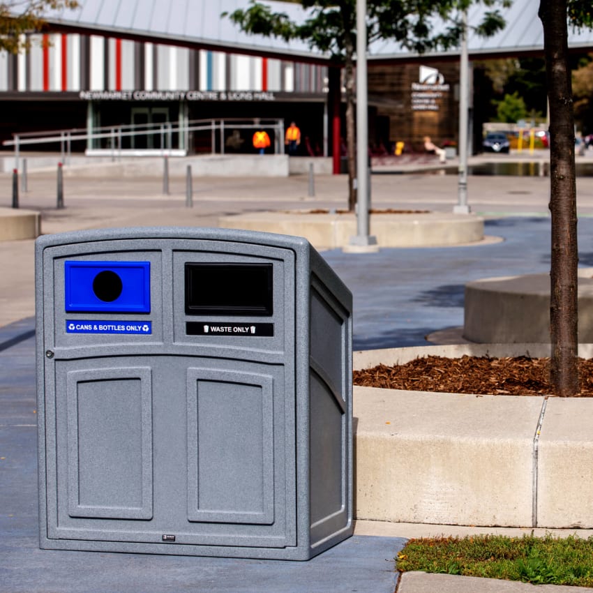 grey plastic recycling and waste bin with sloped top and front hinged door sitting outside at a municipal park
