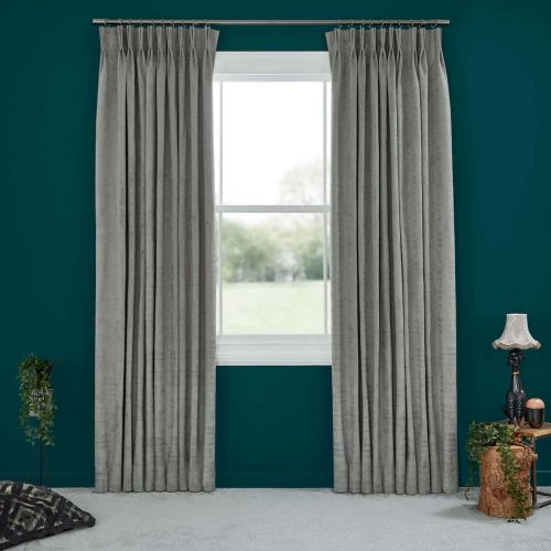 Grey pleated floor-length curtains on a metal rod, flanking and drawn aside to reveal a bright double-hung window, filtering soft daylight in a teal-walled room with carpet, lamp, and plants.
