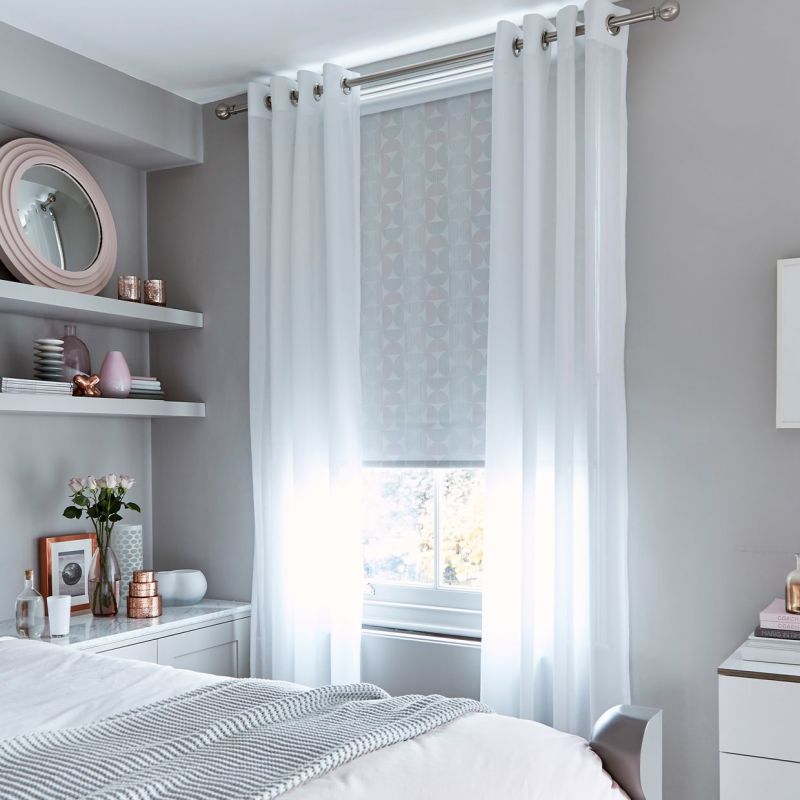 Sheer white grommet-top curtains, layered over a patterned roller blind, hang drawn at a metal rod, softly filtering daylight into a pale-gray bedroom with bedside shelves and soft decor.