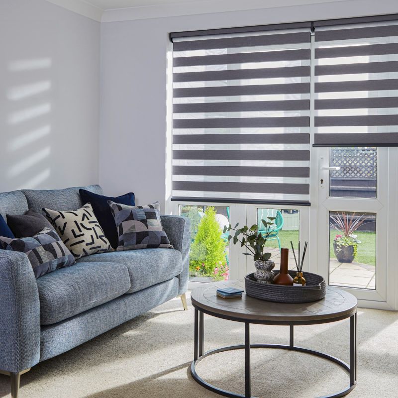 Dual-layer roller blinds with alternating opaque and sheer horizontal stripes, lowered over patio doors and windows, filtering sunlight; living room with blue sofa, patterned cushions, coffee table, garden view.