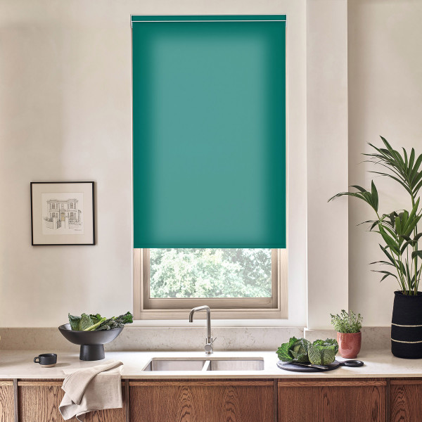 Teal roller blind covers most of a kitchen window above a faucet and sink, countertop holding a bowl of leafy greens, potted plants, and a framed sketch on the wall.