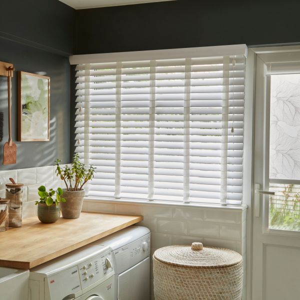 White horizontal slatted blinds cover a wide window, half-closed and filtering daylight; above a wooden counter with plants atop a washer/dryer, woven laundry basket nearby in a laundry room.