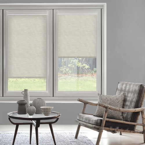 Light-beige fabric roller blinds installed inside the twin window frames, left almost closed and right halfway down, partially blocking light; living room with gray walls, rattan armchair and coffee table.