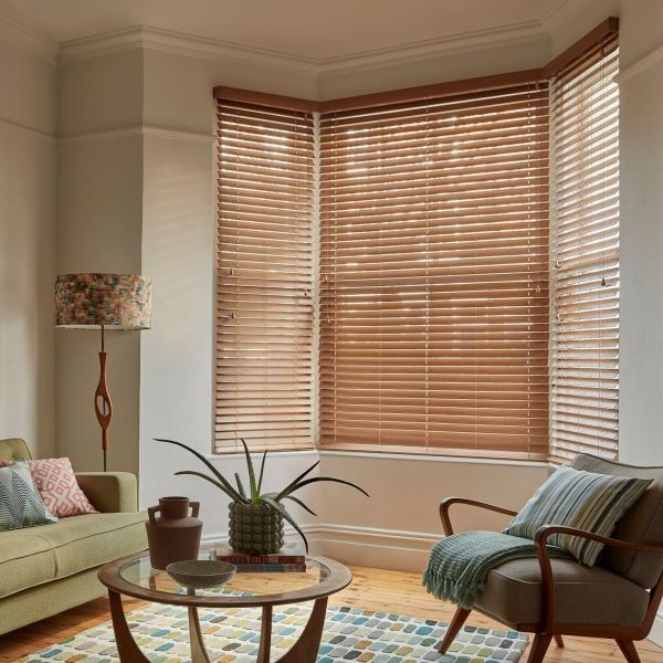 Wooden Venetian blinds covering a three-pane bay window, slats mostly closed and softly filtering daylight; cozy living room with mid-century armchair, sofa, patterned rug, glass coffee table and lamp.