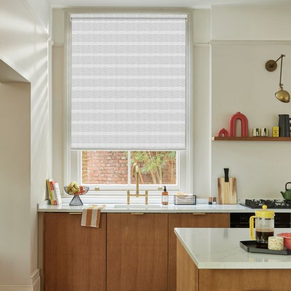 A white textured roller blind, fully lowered across a sash window, diffusing daylight over a kitchen sink with brass faucet, marble countertop, wooden cabinets, open shelf and brick courtyard view.