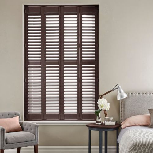 Wooden plantation shutters fill the recessed window, narrow panels with adjustable louvers closed but angled to filter daylight; set in a neutral bedroom corner with bed, side table and armchair.