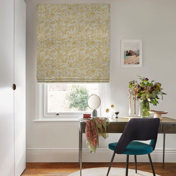 Floral-patterned Roman blind (yellow on cream), drawn closed and softly filtering daylight across a home office desk with chair, vase of flowers, framed coastal print, and neutral-painted room.