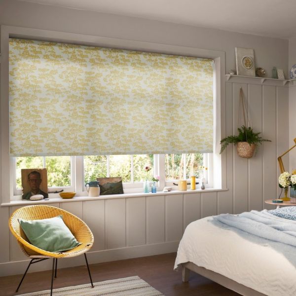 A yellow-floral roller blind lowered over a multi-pane window, filtering soft daylight into a cozy paneled bedroom with a wicker chair, hanging plant, windowsill ornaments and a bed.