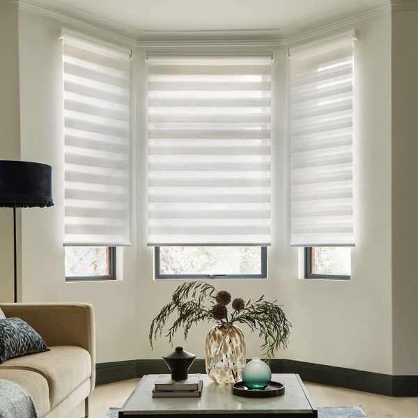 Three white zebra roller blinds lowered across a bay of windows, alternating sheer and opaque bands, filtering soft daylight into a living room with sofa, lamp and coffee table.