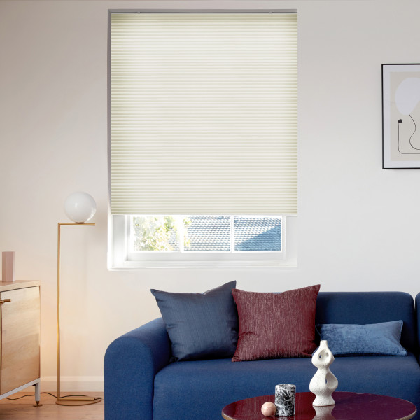 Cream cellular blind (horizontal pleats) covering the window, lowered and gently filtering daylight; living room with blue sofa and cushions, brass floor lamp, wooden sideboard and framed abstract art.