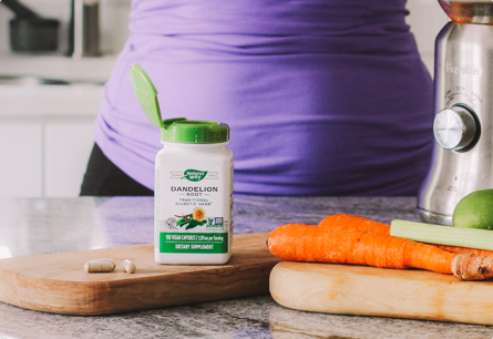 A bottle of Dandelion Root capsules sitting on a cutting board next to carrots and celery.