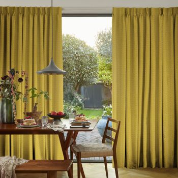 Floor-length mustard-yellow pleated curtains, drawn aside in gathered folds, frame a glass sliding door, admitting soft daylight onto a dining table with pancakes and a lush backyard garden.
