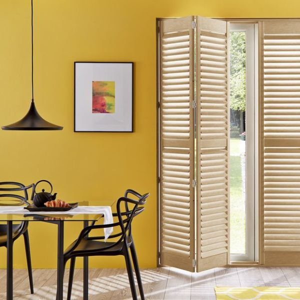 Wooden plantation shutters folded open to the right, bi-fold panels with angled louvers letting daylight through. In a yellow-walled dining area with black pendant lamp, table, chairs, and garden view.