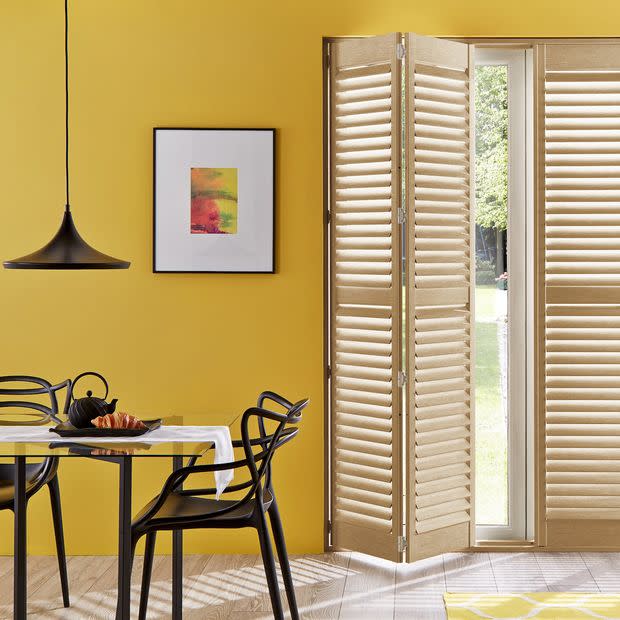 Wooden plantation shutters folded open to the right, bi-fold panels with angled louvers letting daylight through. In a yellow-walled dining area with black pendant lamp, table, chairs, and garden view.
