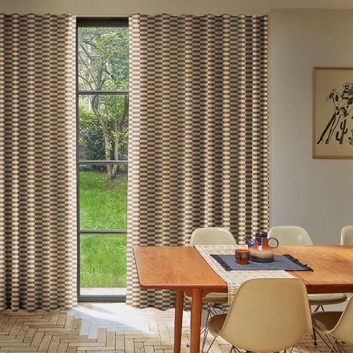 Floor-to-ceiling checkered brown-and-cream pleated curtains, drawn aside to reveal a narrow four-pane window, filtering daylight; a sunlit dining room with wooden table, beige chairs, lawn and tree outside.