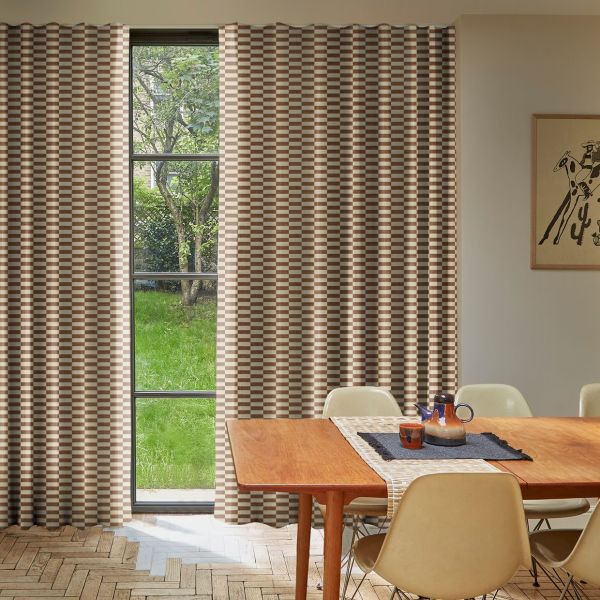 Floor-to-ceiling checkered brown-and-cream pleated curtains, drawn aside to reveal a narrow four-pane window, filtering daylight; a sunlit dining room with wooden table, beige chairs, lawn and tree outside.
