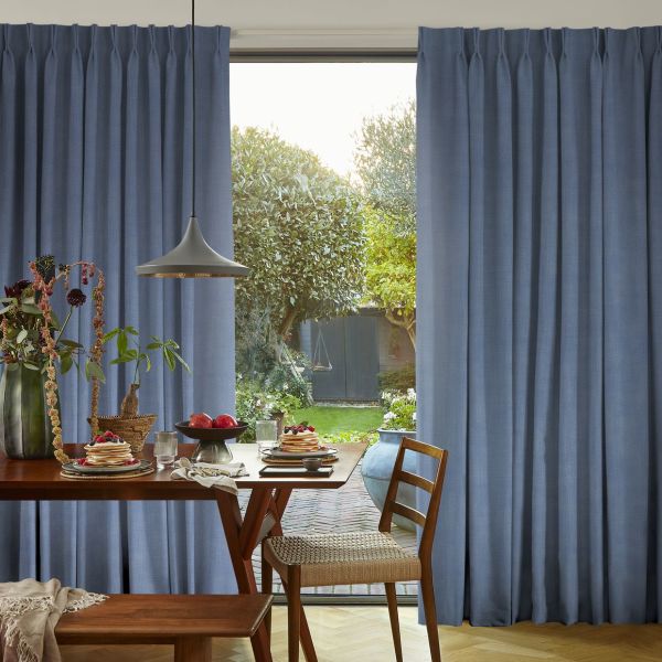 Blue floor-length pleated curtains, hung and parted centrally to reveal sunlight; framing a wooden dining table laid for breakfast in a softly lit room that opens onto a green backyard.