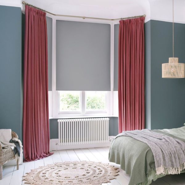 Floor-length pleated mauve curtains flank a lowered grey roller blind, framing and shading a bay window in a teal-walled bedroom with white floors, radiator, bed, and greenery visible outside.
