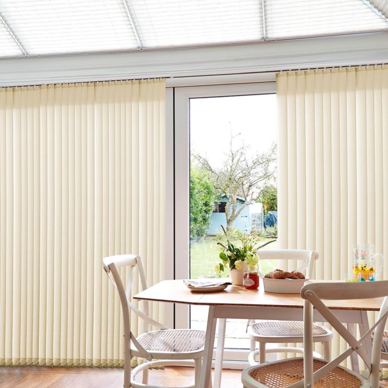 Beige vertical blinds over a patio door, parted centrally, filtering daylight. Outside a garden with a tree and shed. Inside a bright dining area with white chairs, table, vase.