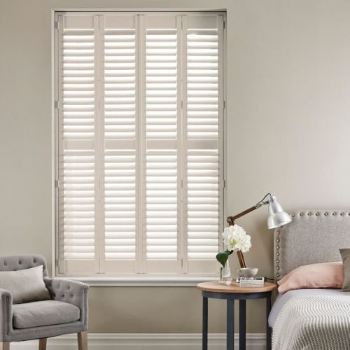 White full-height plantation shutters covering a tall window, louvers tilted to filter soft daylight; set in a neutral-toned bedroom with upholstered bed, bedside table lamp, vase of flowers and armchair.