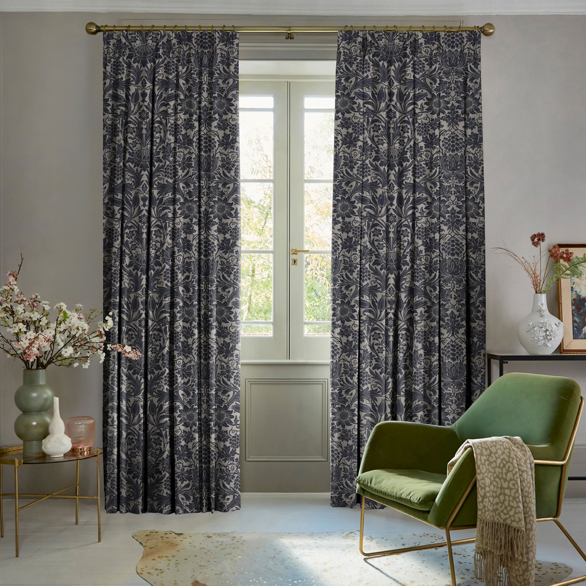 Patterned navy-grey floral drapery panels on a brass rod, drawn partly aside to frame a tall paneled French window, filtering soft daylight in a neutral, furnished sitting room.