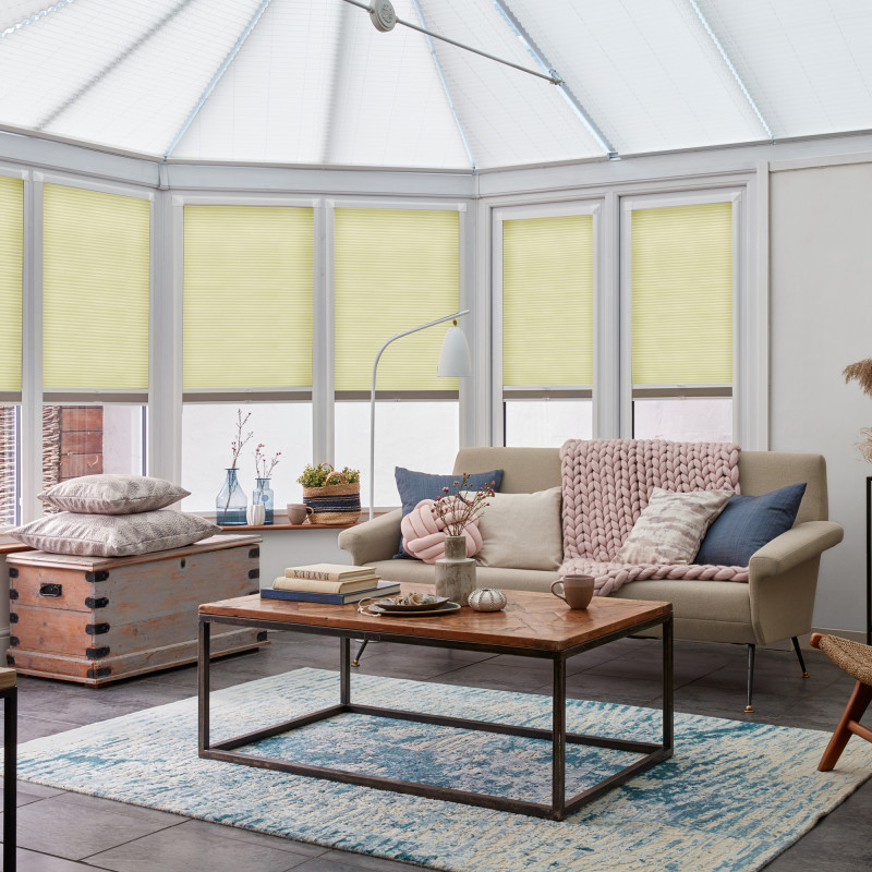 Light-yellow pleated cellular blinds covering bay windows and roof panels, partially lowered and filtering diffuse daylight, in a bright conservatory-style living room with sofa, coffee table, and pastel decor.